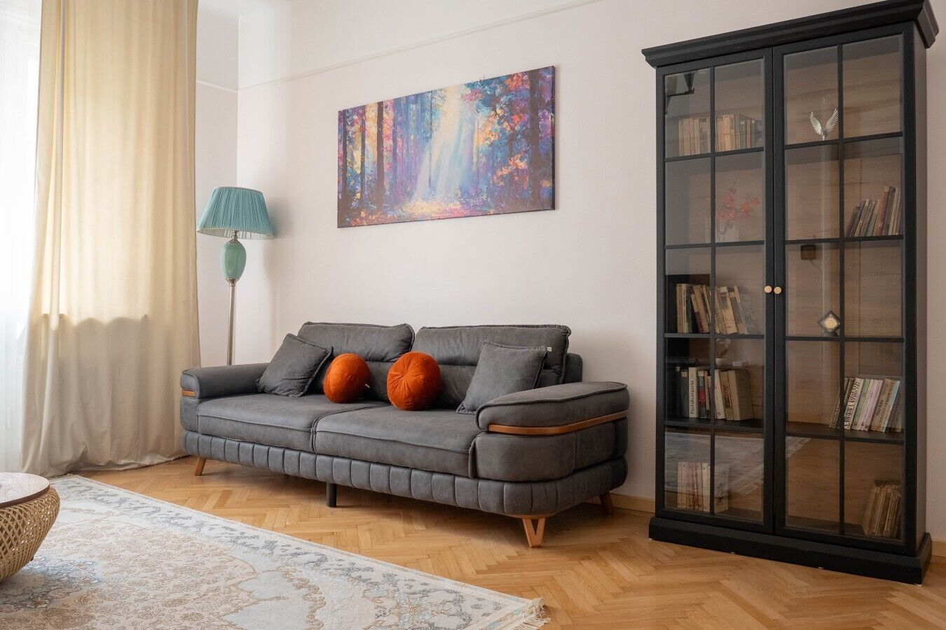 Un salon avec un canapé gris, des coussins orange, une lampe sur pied, une bibliothèque et un tableau accroché au mur.