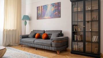 Un salon avec un canapé gris, des coussins orange, une lampe sur pied, une bibliothèque et un tableau accroché au mur.