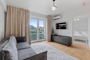 Ein modernes Wohnzimmer mit einer Couch, einem an der Wand befestigten Fernseher und einem Balkon mit Blick auf Gebäude.