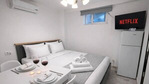 Ein kleiner, moderner Raum mit einem Bett, einem für zwei Personen gedeckten Tisch mit Weingläsern, einem Fernseher an der Wand und einem Kühlschrank.