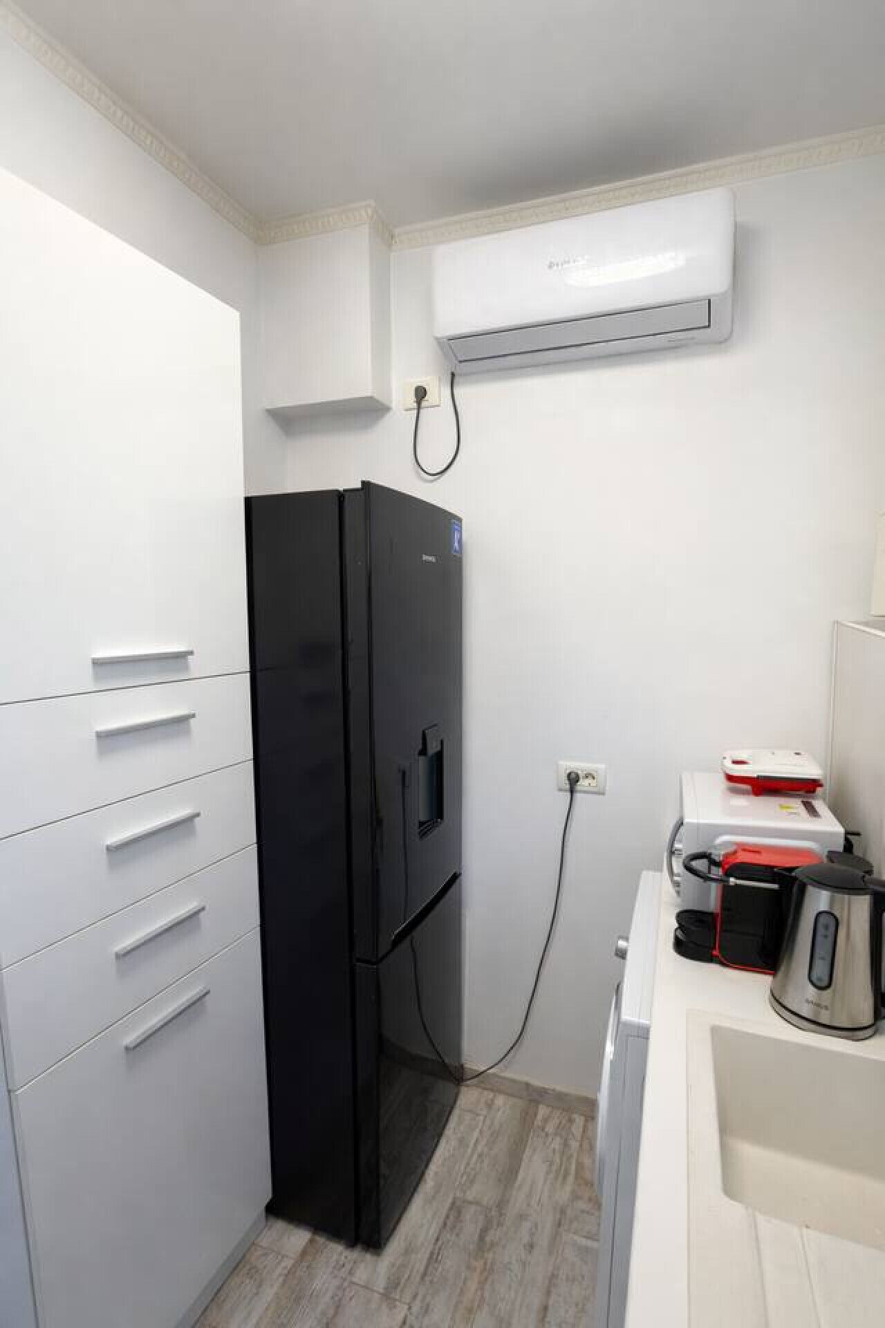 A compact kitchen with a black refrigerator, white cabinetry, a coffee maker, and an air conditioning unit.