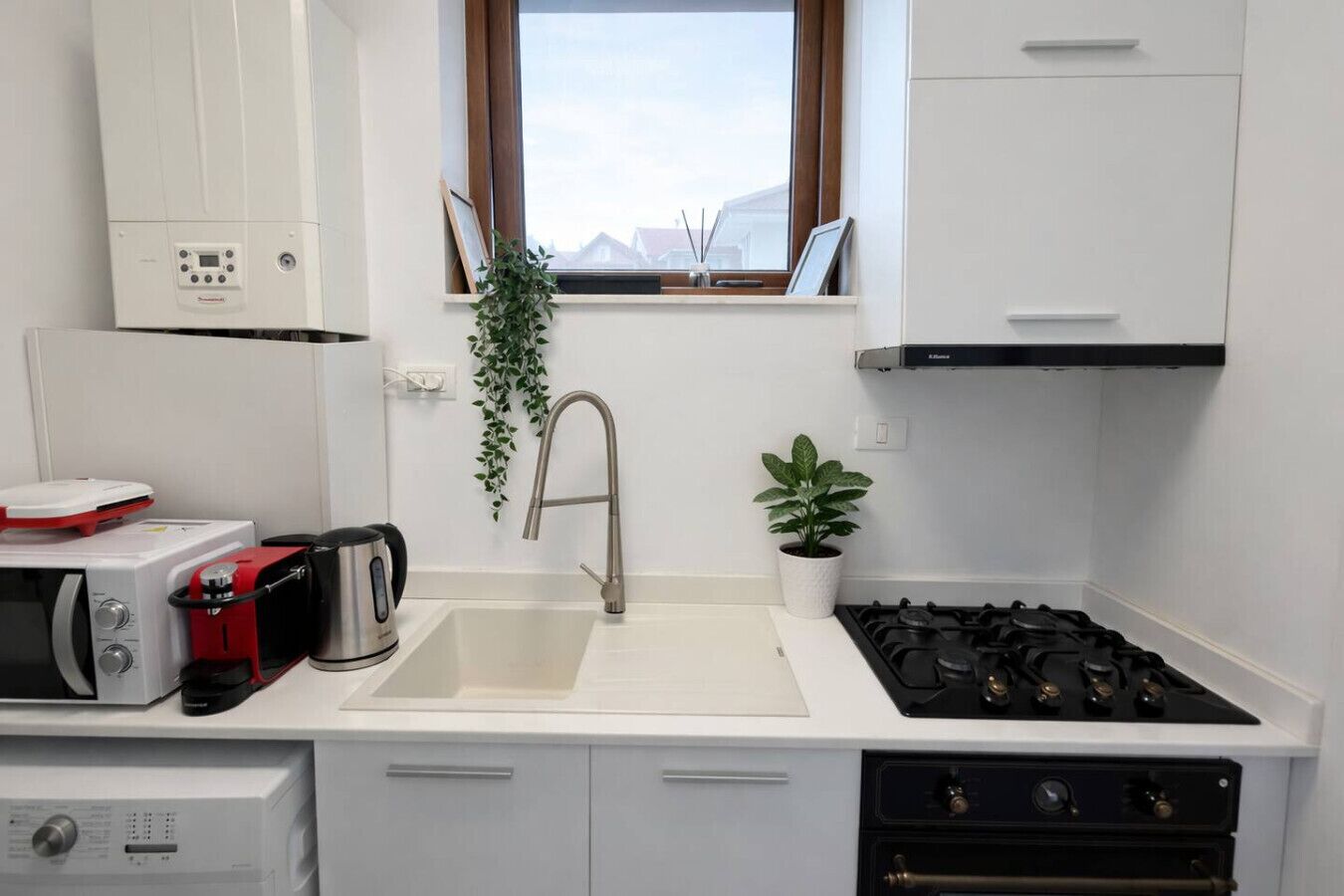 Une cuisine compacte avec un plan de travail blanc, un évier en acier inoxydable, une cuisinière à gaz, un four à micro-ondes et une petite plante en pot.