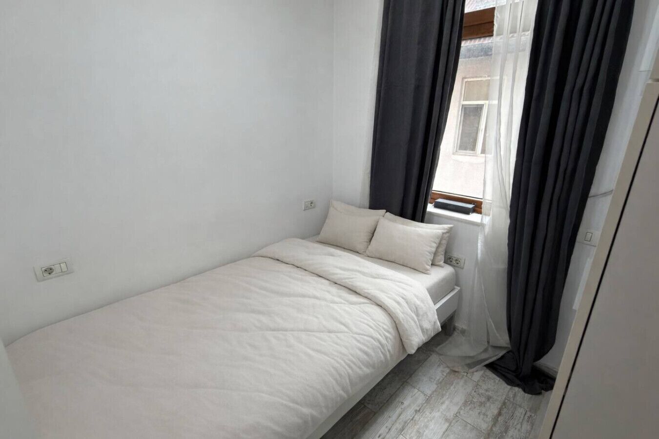 Een kleine kamer met een enkel bed, een raam en een stopcontact aan de muur.