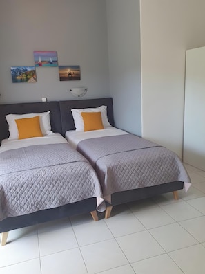 A double bed with a grey quilted bedspread and two pillows, one white and one mustard yellow, in a room with a grey wall and a white floor.