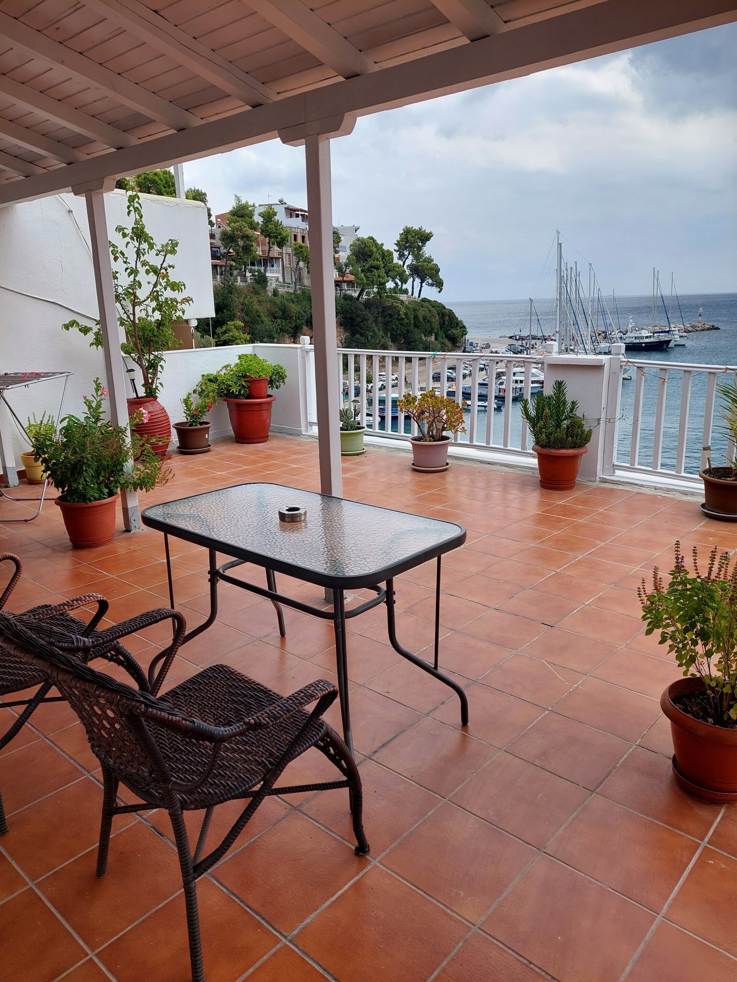 A terrace with a table and chairs, potted plants, and a view of the sea and boats.
