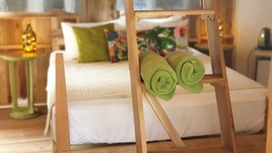 A room with a bed, a wooden stand with rolled towels, a small table with a lamp, and a window with curtains.