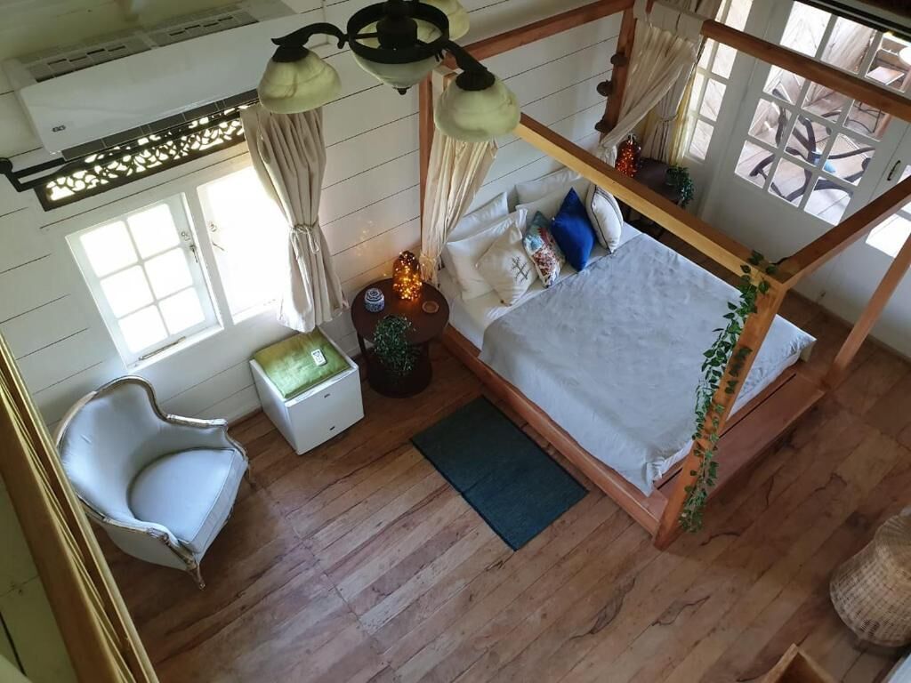 A wooden four-poster bed with a white bedspread, a white chair, a small bedside table, and a window with curtains.