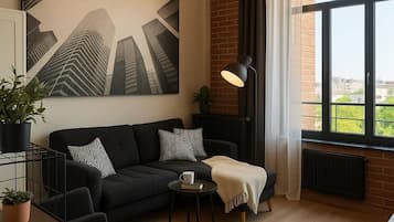 Un salon moderne avec un canapé noir, une table basse, un lampadaire et une grande fenêtre donnant sur des immeubles.