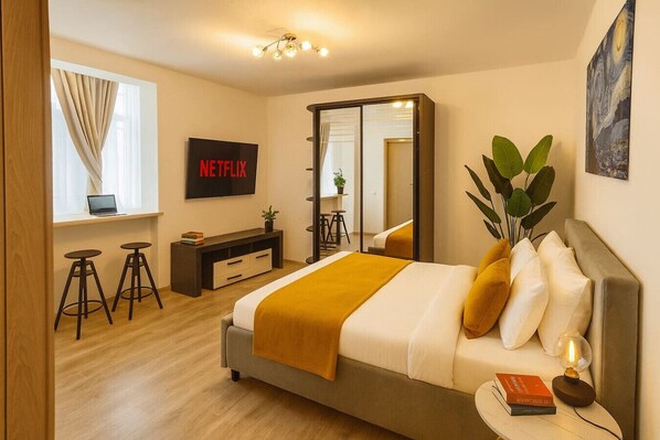 A modern hotel room with a bed, a desk with a laptop, a mirror, a plant, and a wall-mounted TV. - Stylish & Comfortable Studio Retreat (Bucharest)