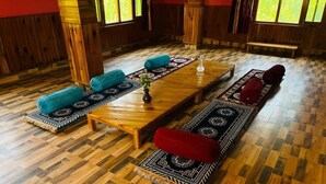 A room with a long wooden table, blue and red cushions, and patterned rugs. - Applewood Heaven (Bhuntar)
