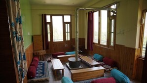 A room with wooden walls, large windows, a central heating system, and multiple seating areas with cushions. - Applewood Heaven (Bhuntar)