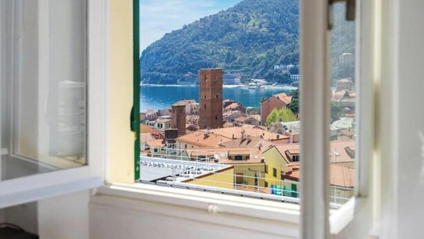 A window view of a coastal town with buildings, a prominent tower, and a hillside. - Hostdomus - Esther House (Noli)