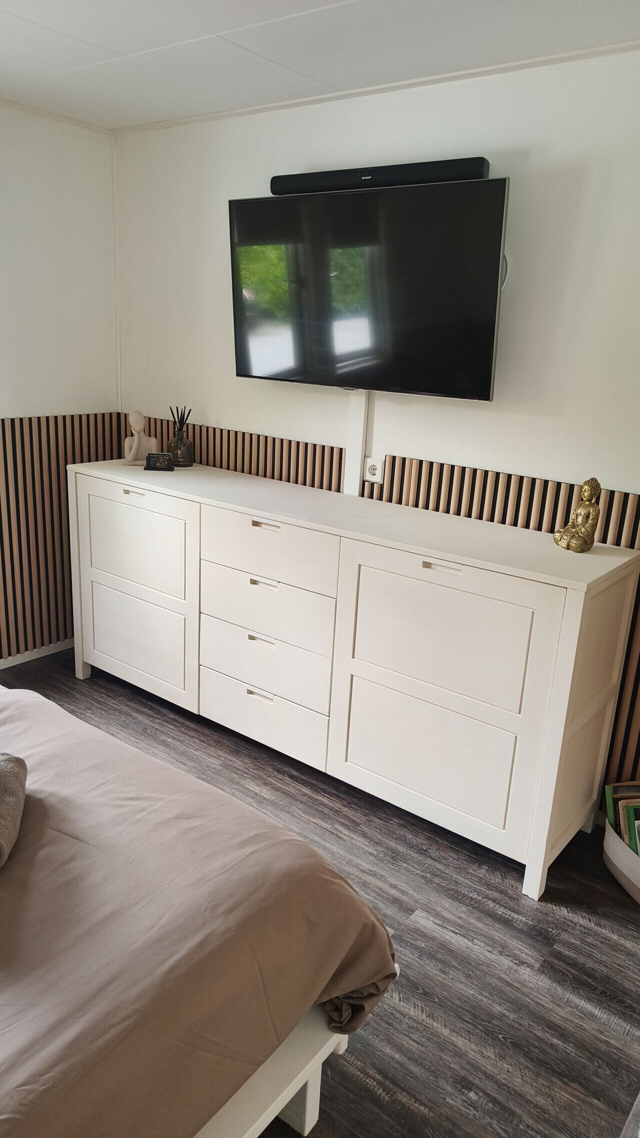 A bedroom with a bed, a white dresser, a flat-screen TV mounted above it, and a small Buddha statue on the dresser.