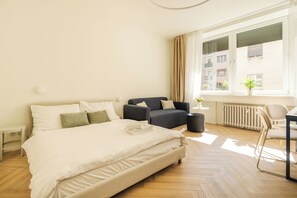 A bedroom with a bed, two sofas, a chair, a small table, a potted plant, and a window with a view of buildings.