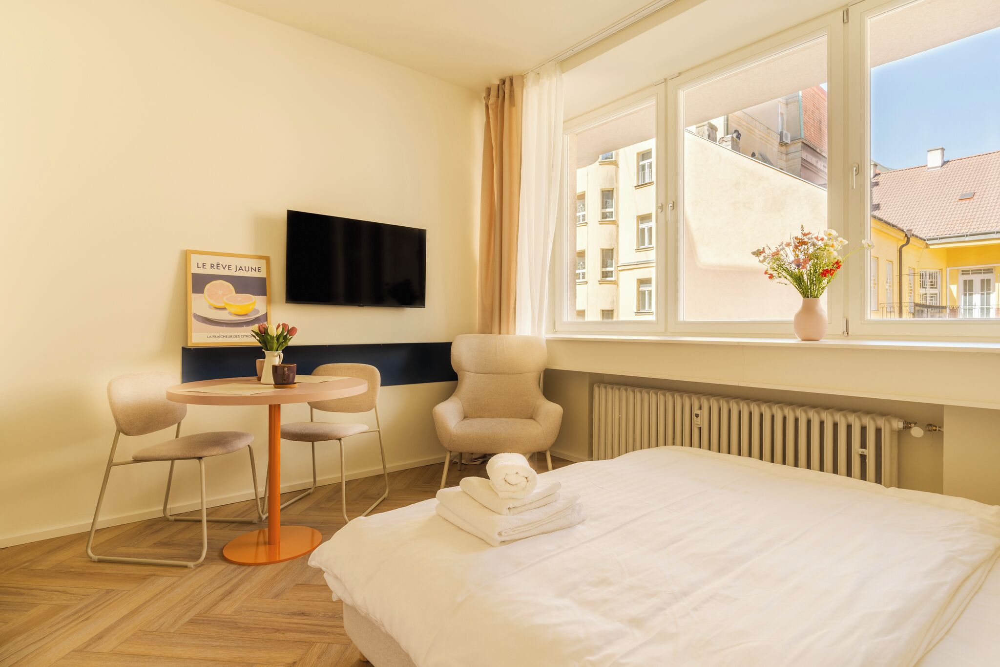 Hotellihuone, jossa on sänky, pyöreä pöytä tuoleineen, televisio, ikkuna, josta on näkymä rakennuksiin, ja juliste seinällä.