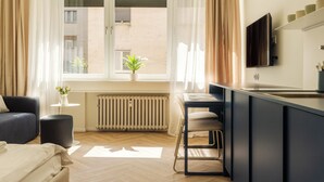 A modern hotel room with a sofa, a dining table with chairs, a TV mounted on the wall, and a window with a view of the outside. - LOCA by Ambiente Apartments (Bratislava)