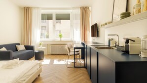 A modern hotel room with a sofa, a dining table, a kitchen area with a coffee machine, and a window with curtains. - LOCA by Ambiente Apartments (Bratislava)