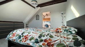 A bedroom with a sloped ceiling, a bed with a leaf-patterned bedspread, a bedside lamp, and a view into an adjacent room. - Appartement Cozy et design, position centrale, complètement équipé. (Perwez)
