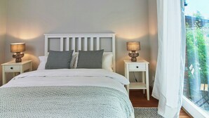 A bed with a white headboard, two bedside tables with lamps, patterned pillows, and a view of greenery through a window. - Artist apartment at Aukrug nature park (Meezen)
