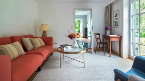 A living room with a sofa, a coffee table, a lamp, a vase with flowers, a chair, a desk, and a window with a view. - Artist apartment at Aukrug nature park (Meezen)