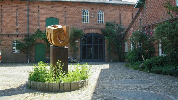 A brick building with a large arched entrance, a wooden sculpture, and a cobblestone courtyard. - Artist apartment at Aukrug nature park (Meezen)