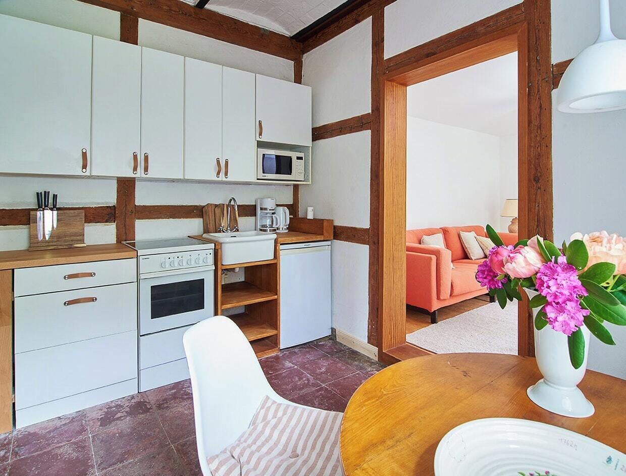 A compact kitchen with white cabinets, a microwave, a coffee maker, and a small refrigerator. There is a dining table with a white chair and a vase of flowers.