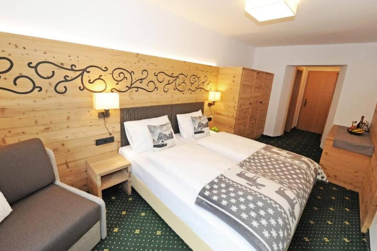 A hotel room with a wooden headboard, a bed with white bedding, a nightstand, a sofa, and a wooden wardrobe.