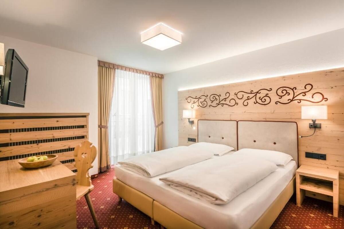 A hotel room with a double bed, a wooden headboard with decorative lights, a small bedside table, a desk with a chair and a bowl of fruit, and a large window with curtains.