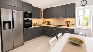 Une cuisine moderne avec des meubles de couleur sombre, des appareils en acier inoxydable, un plan de travail en bois et un coin repas comprenant une table et des chaises blanches.