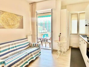 Een kamer met een gestreept bankje, een kleine eettafel met stoelen, een witte stoel, en een keukenhoek met kastjes en een fornuis.