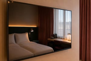 Quarto de hotel com cama, luminária de cabeceira, escrivaninha, cadeira e uma janela com cortinas.