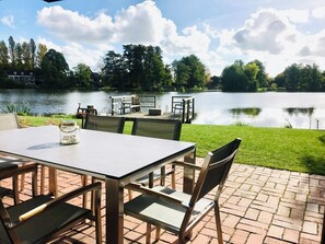 Un patio al aire libre con una mesa y sillas, una terraza de madera y vistas a un lago con árboles y un muelle.