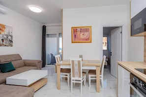 A modern living room with a sofa, a dining table with chairs, a kitchen area with a stove, and a sliding glass door leading to a balcony.