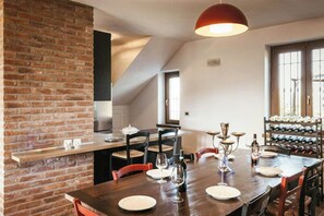 Zona pranzo con parete in mattoni, tavolo in legno, sedie, bicchieri da vino e cantinetta.