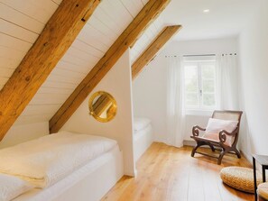 A bedroom with a sloped ceiling, a bed, a chair, a window with curtains, and a round mirror. - Farmhouse comfort in the Uckermark region (Weggun)