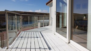 A wooden deck with glass railings, a clear view of the sea, and a building with large windows. - NEW to VRBO: Spacious House w/ Coastal Views & Private Garden (Whiteness)