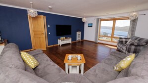 A living room with a sofa set, a coffee table, a TV, and a window with a view. - NEW to VRBO: Spacious House w/ Coastal Views & Private Garden (Whiteness)