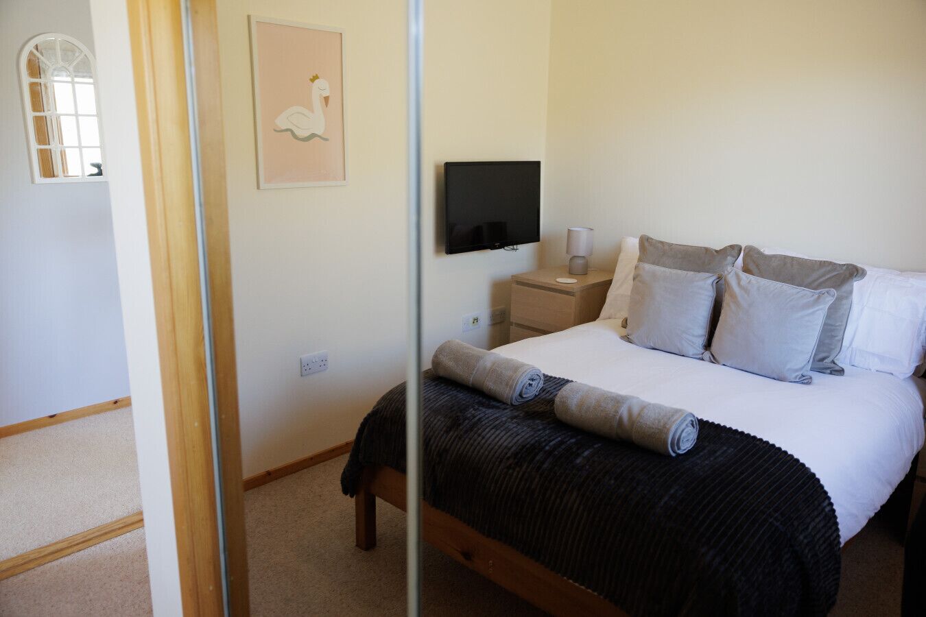 A bedroom with a bed, pillows, a bedside table with a lamp, a TV mounted on the wall, and a framed picture.