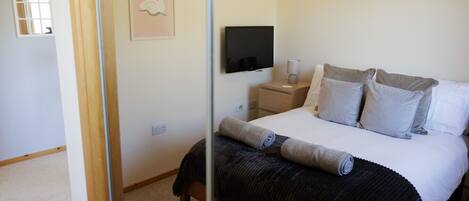 A bedroom with a bed, pillows, a bedside table with a lamp, a TV mounted on the wall, and a framed picture.