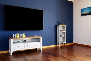 Una habitación con una televisión de pantalla plana grande montada en la pared, un mueble blanco con la palabra 'HOME' y una estantería blanca, sobre un piso de madera.