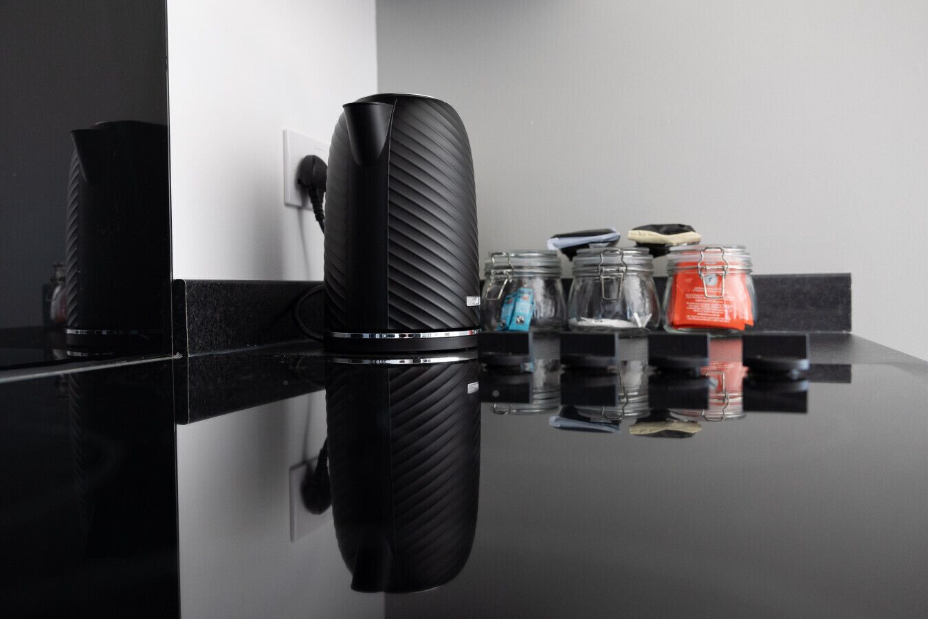 A black, ribbed, cylindrical object on a reflective surface, a power outlet, and a shelf with jars and containers.