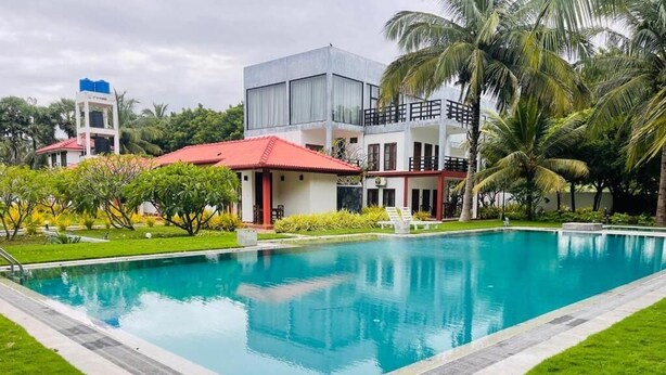 A swimming pool with a surrounding grassy area, a building with a red roof, and palm trees. - NN Beach Resort Nilaveli (Trincomalee)