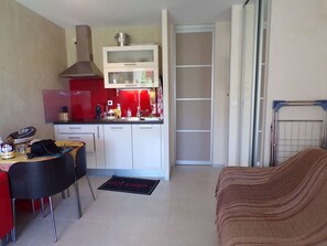 A compact kitchen with a dining area, a red chair, a black table, and a brown sofa. - Cozy Studio with Pool, Near Beach & Shops, South-Facing Garden Terrace (La Grande-Motte)