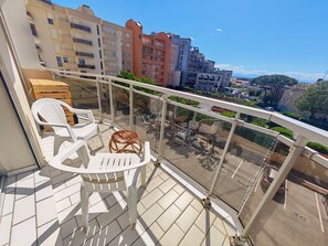 Ein Balkon mit weißen Plastikstühlen und einem Korbstuhl-Tisch, mit Blick auf eine Stadtlandschaft mit verschiedenen Gebäuden und wolkenlosem Himmel.