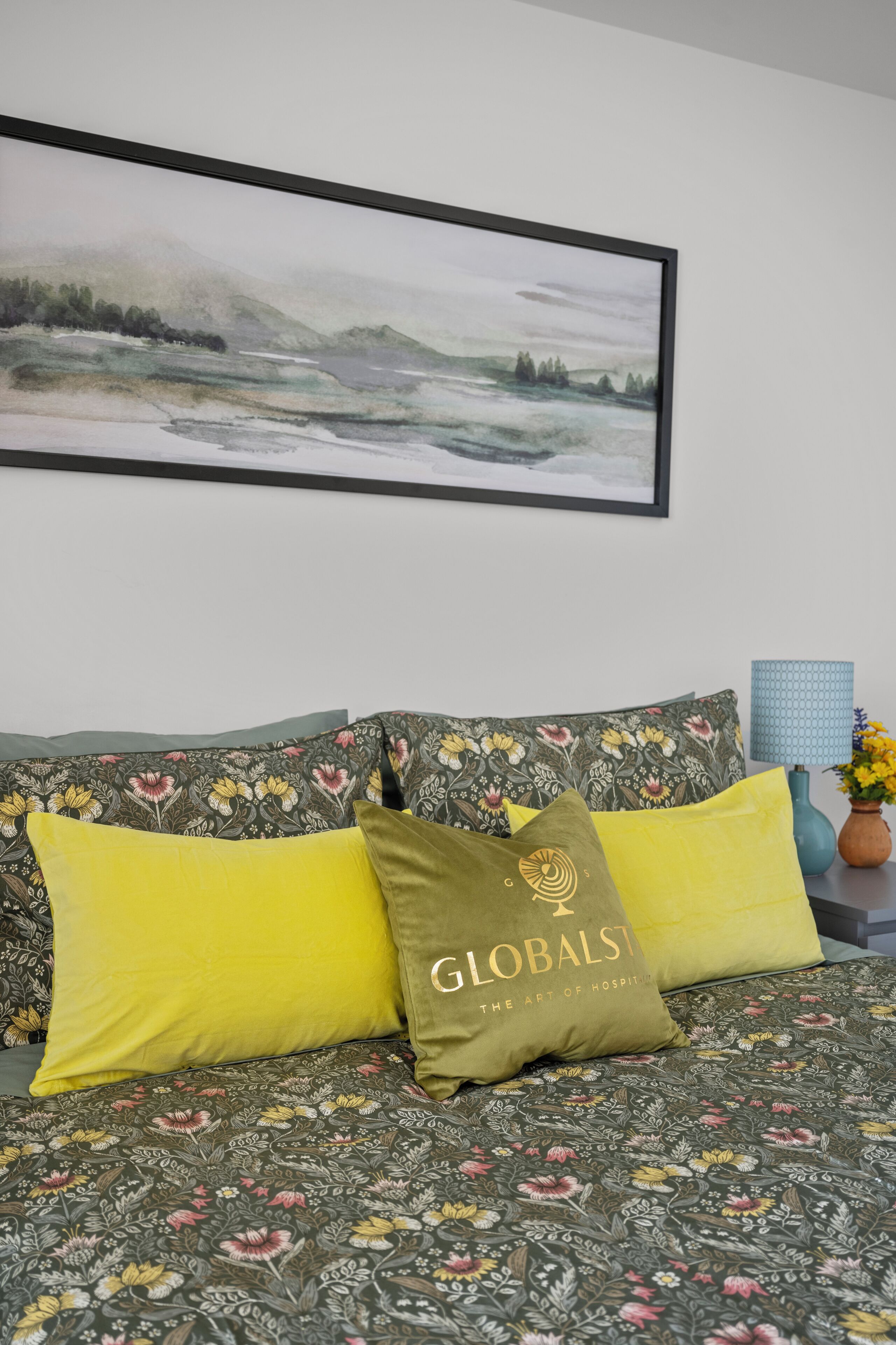Un lit avec une literie à motifs floraux, un tableau représentant un paysage encadré et un vase bleu contenant des fleurs jaunes.