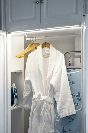 Ein weißer Bademantel hängt an einem Holzkleiderbügel in einem Badezimmer mit einem blauen Handtuchhalter und einem weißen Schrank mit integriertem Bügeleisen.