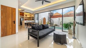 A modern living room with a black sofa, a round wooden coffee table, a ceiling fan, and a large window offering a view of a pool and outdoor area. - Alisa 3 Villa (Kerobokan)