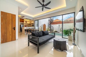 Ein modernes Wohnzimmer mit einer schwarzen Couch, einem runden Holztisch, einem Deckenventilator und einem großen Fenster mit Blick auf Pool und Außenbereich.