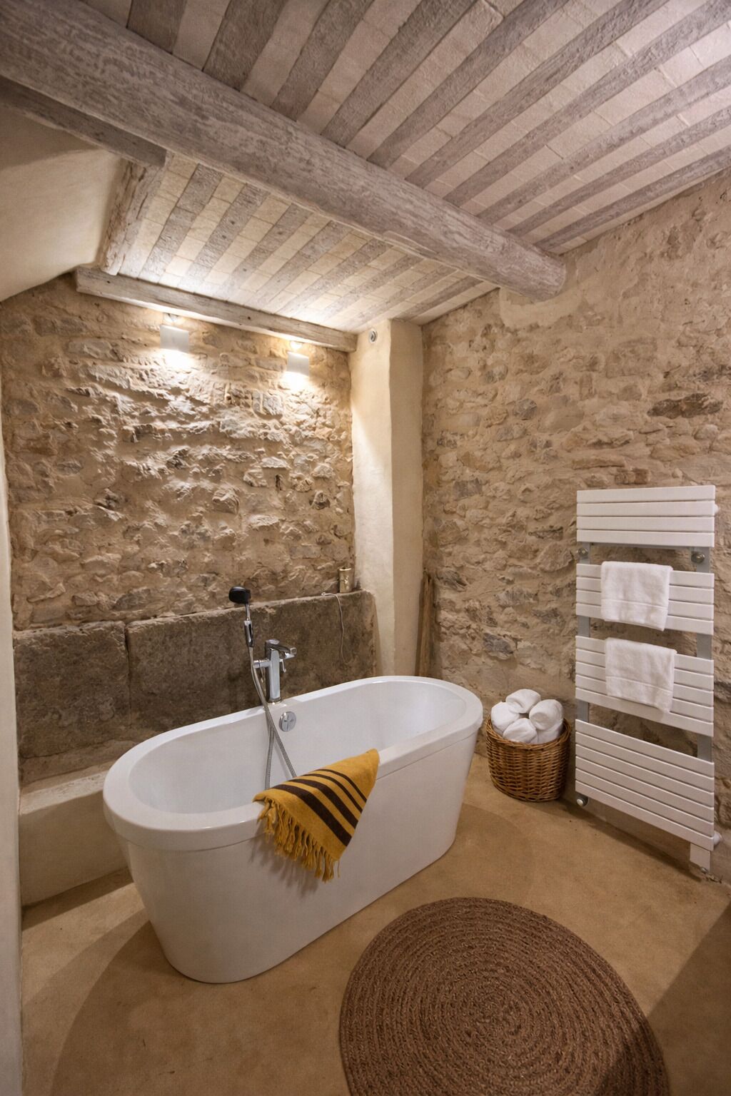 A stone-walled bathroom with a freestanding bathtub, a towel rack, and a woven mat.