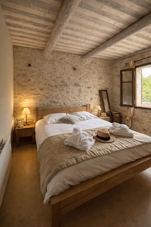 Ein Schlafzimmer mit einem hölzernen Bett, Steinwänden, einem Nachttisch mit Lampe und einem Fenster mit Vorhängen.